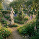 Goddess of Harvest Marble Sculpture by Giant Sculptures, 160cm female figure with floral crown on a stone plinth in a cottage garden with foxgloves and roses.