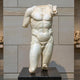 The Classical Male Marble Torso Sculpture - 130cm by Giant Sculptures features a muscular figure in natural white marble, draped over one shoulder and set on a black pedestal against a stone wall.