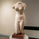 The Female V2 Marble Torso Sculpture - 130cm by Giant Sculptures, a large headless and armless female torso missing part of the legs, is displayed on a reddish-brown stone base in a museum setting.