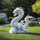 The Mystical Dragon White Marble Outdoor Sculpture by Giant Sculptures features a 100cm serpentine-bodied dragon with a fierce expression, detailed scales, and sharp claws. Set on garden grass, lush greenery and rocks in the background enhance its serene atmosphere.