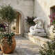 A Mediterranean-style courtyard features the Coiled Snake White Marble Outdoor Sculpture (100cm) by Giant Sculptures in a fountain, surrounded by potted plants, purple flowers, and rustic stone walls with soft sunlight filtering in.