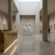 A modern art gallery with minimalist design showcases the Ferocious Snake White Marble Outdoor Sculpture – 100cm by Giant Sculptures as its centerpiece. Marble floors and high ceilings create a spacious, light-filled space as a visitor admires nearby framed art.