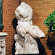 Seated Male Marble Torso Sculpture in natural white marble displayed against a brick wall with green foliage, showing muscular chest detail and draped waist.