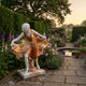 Dancing Woman Marble Sculpture 120cm by Giant Sculptures, white figure lifting a flowing amber-brown skirt, on stone paving beside a garden pond and flowers.