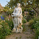 Goddess with Cornucopia Marble Sculpture by Giant Sculptures standing 180cm in a cottage garden path, draped figure cradling a cornucopia among flowers.
