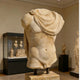 The Draped Marble Torso Sculpture - 120cm by Giant Sculptures, missing its head, arms, and lower legs, is elegantly displayed in a museum gallery among other textured marble artworks.