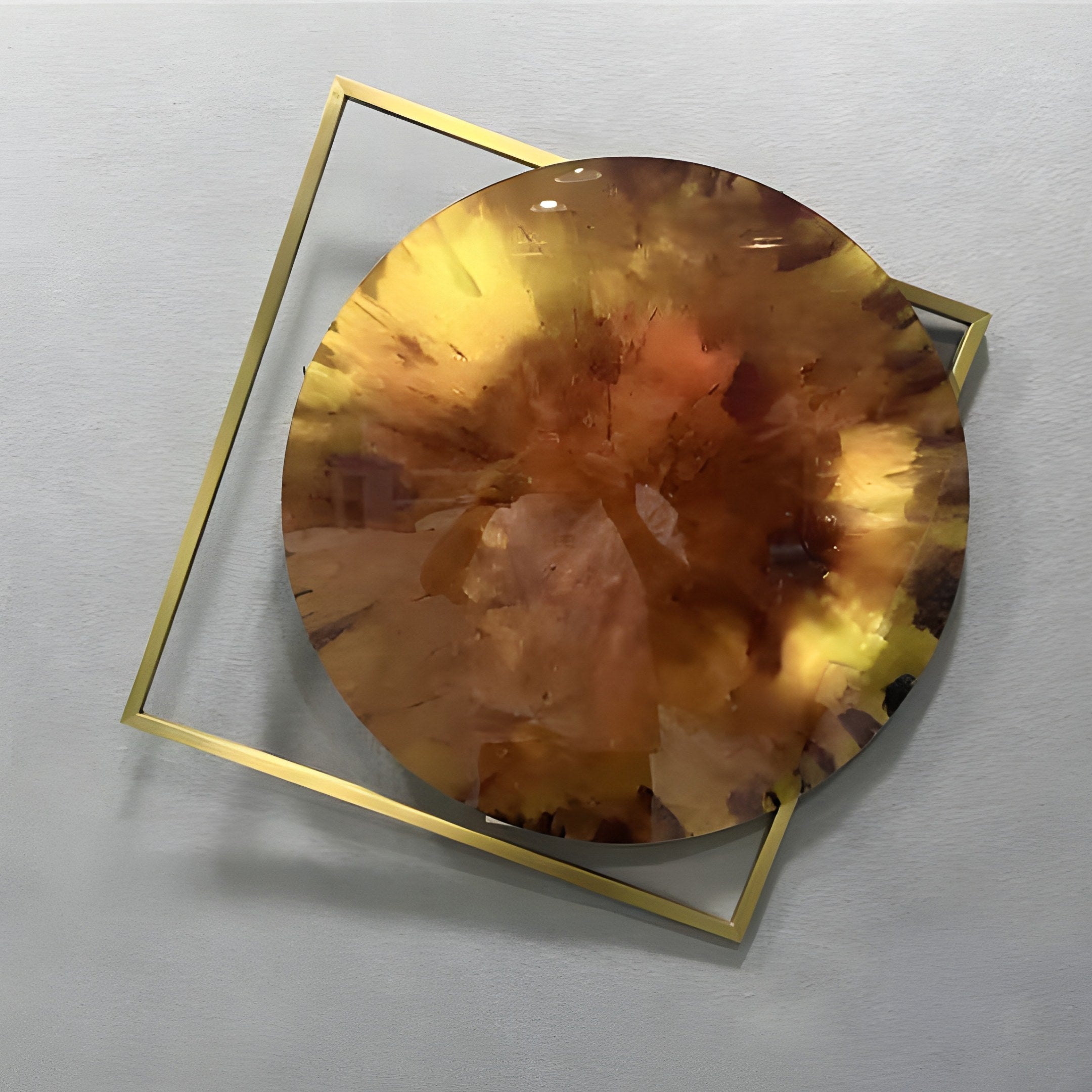Aurenza Amber Acrylic & Metal Frame 3D Wall Art featuring a glossy amber disc with warm sienna and ochre tones mounted over a diagonally angled gold-tone square frame on a light grey wall.
