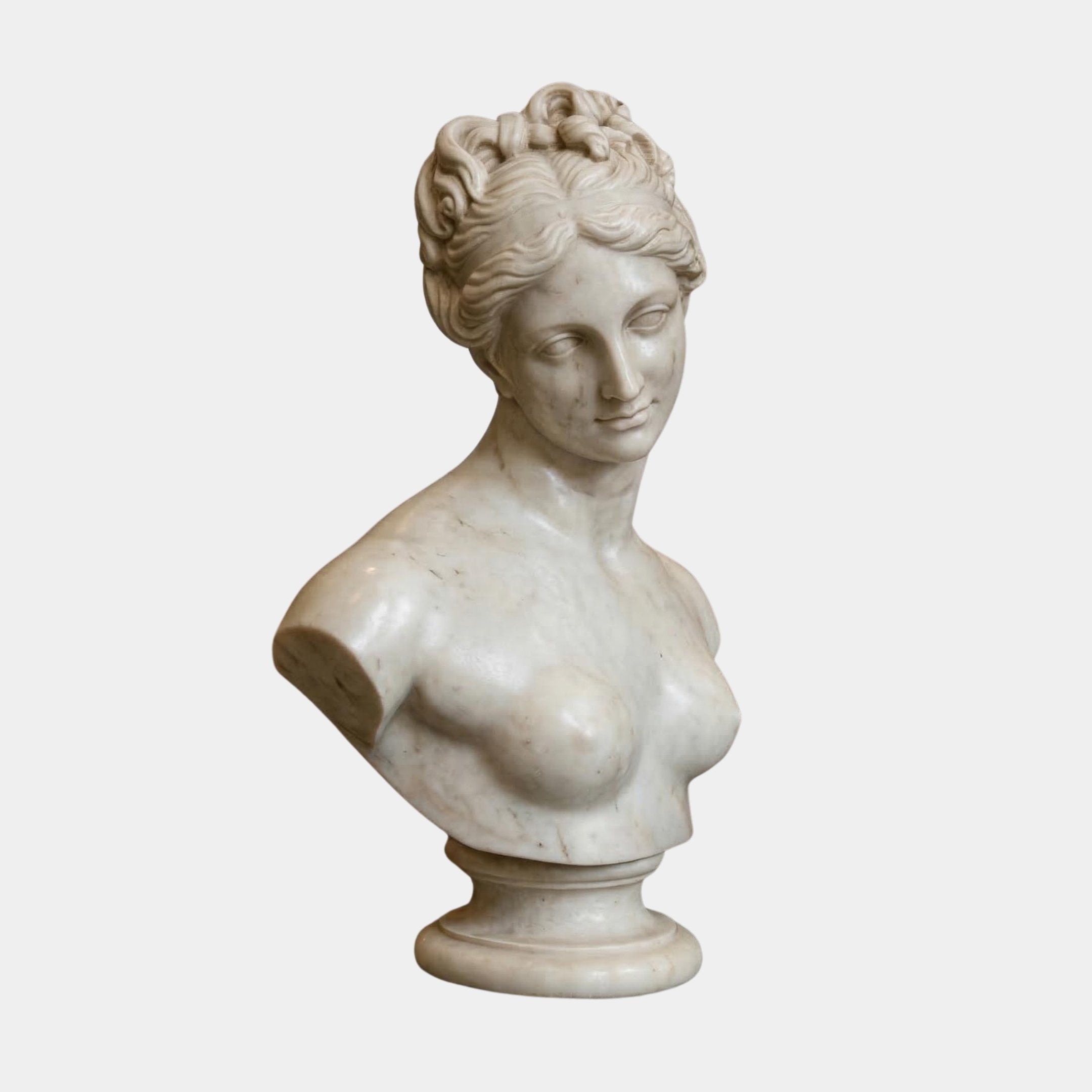 The Aphrodite Marble Bust Sculpture by Giant Sculptures is a 65cm white marble figure with wavy hair pinned up, a serene expression, and draped shoulders, set on a round pedestal base against a plain background.