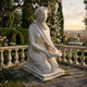 The Veiled Lady Marble Sculpture - 130cm by Giant Sculptures depicts a kneeling white marble figure holding a tray with a sculpted flame, set in an elegant garden with blooms, trees, and distant buildings under a sunset sky.
