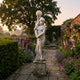 Female Figure Lamp Marble Sculpture by Giant Sculptures on a stone plinth along a flagstone path edged with lavender, foxgloves and roses at dusk.