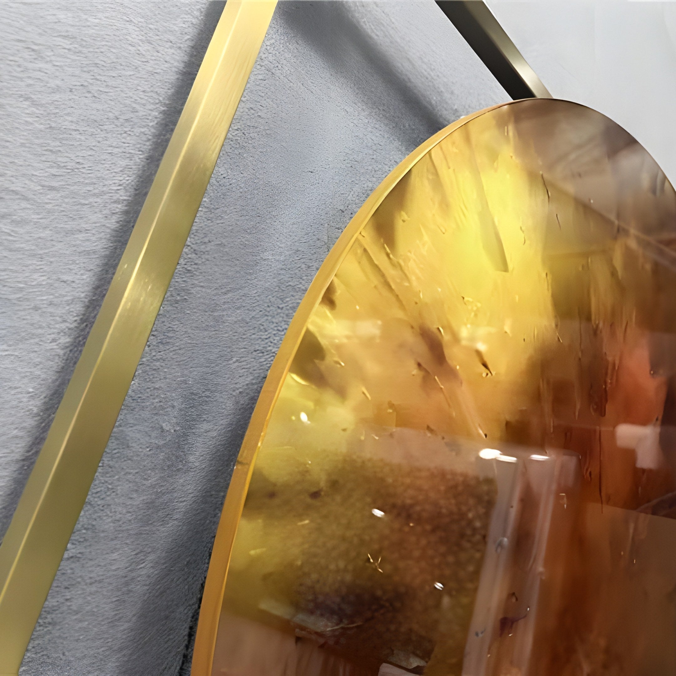 Close-up angled view of the Aurenza Amber Acrylic & Metal Frame 3D Wall Art showing the glossy amber acrylic disc with gold-to-sienna gradient and gold-tone metal frame against a grey textured wall.
