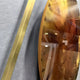 Close-up of the Aurenza Amber Acrylic & Metal Frame 3D Wall Art showing the high-gloss amber acrylic disc and gold-tone metal frame edge mounted on a grey textured wall.