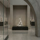 The Giant Coiled Snake White Marble Outdoor Sculpture by Giant Sculptures (160cm) stands on a dark pedestal in the gallery, framed by an arched stone structure and surrounded by glass cases and classical statues, showcasing its impressive artistry.
