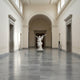 The Triumphant Trumpet Angel Marble Outdoor Sculpture (190cm) by Giant Sculptures, with outstretched arms, stands in the center of a spacious museum hall featuring high ceilings, arched doorways, and polished gray marble floors.