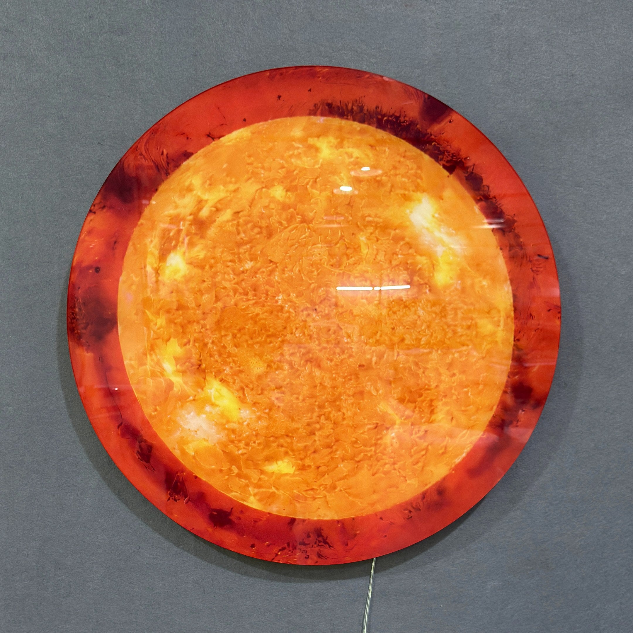 Galaxia Sun Crystal Porcelain LED Wall Art by Giant Sculptures mounted on a grey wall, displaying a glowing circular sun design in amber, orange, and deep red tones with a visible power cord.