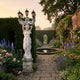 Classical Woman Marble Lamp Sculpture 300cm by Giant Sculptures, draped figure holding a five-lantern candelabra on a carved pedestal in a walled flower garden at dusk.