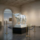 The Majestic Bull White Marble Outdoor Sculpture (120cm) by Giant Sculptures is displayed on a pedestal in a spacious gallery, surrounded by a blue rope barrier, with marble statues and framed paintings in the background.