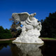 Amid clouds in a clear blue sky, the 280cm Flying Pegasus White Marble Outdoor Sculpture by Giant Sculptures rears on its hind legs over a tranquil pond, surrounded by lush green trees.