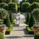 The Robust Bull White Marble Outdoor Sculpture - 120cm by Giant Sculptures stands on a stone pedestal amid potted plants, topiary trees, and gravel paths, with lush green trees in the background of this elegant formal garden display.