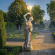 Dancing Lady Lamp Marble Sculpture 180cm by Giant Sculptures, a draped female figure holding an illuminated globe lamp aloft, displayed on a stone base in a formal hedged garden.