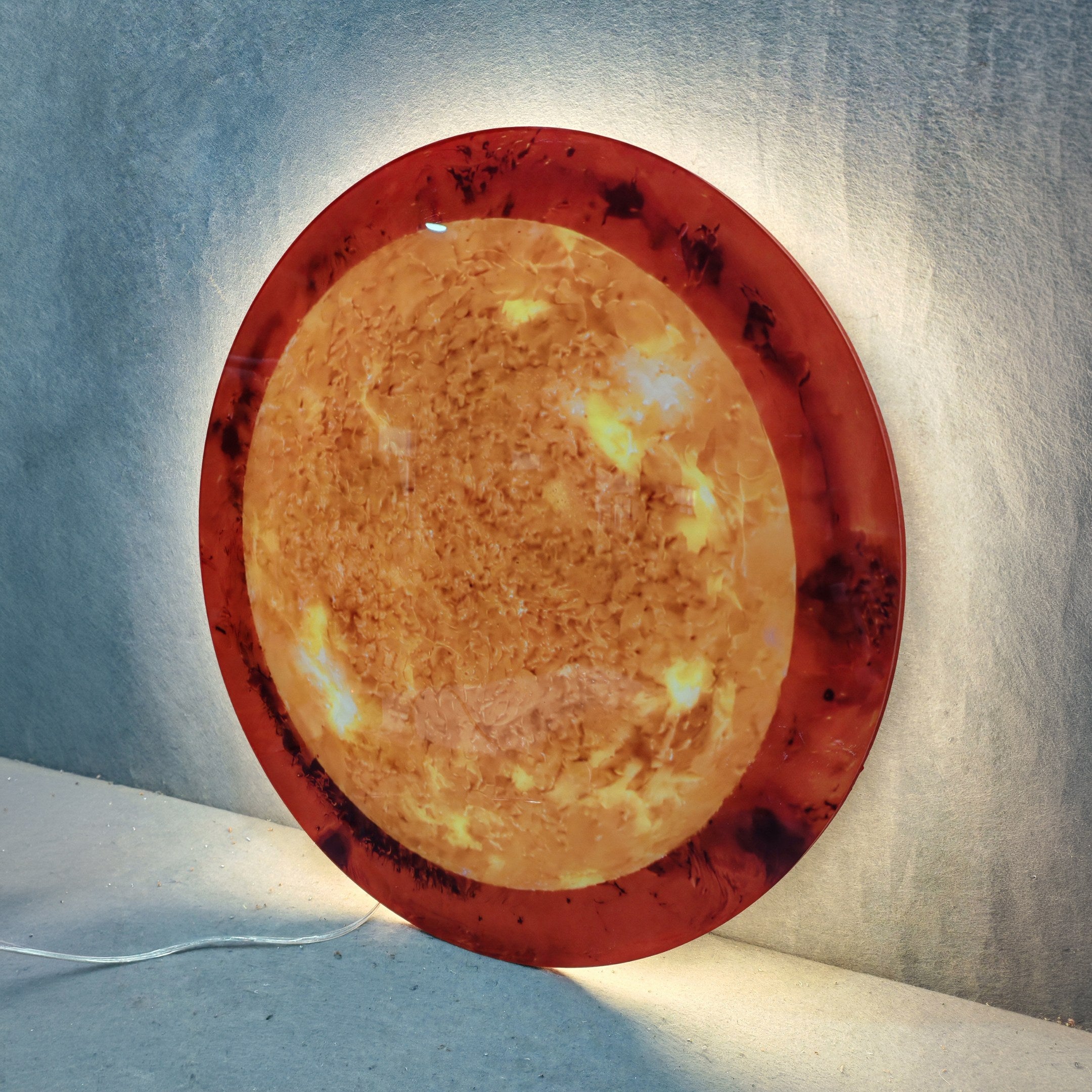 Galaxia Sun Crystal Porcelain LED Wall Art by Giant Sculptures displaying a round amber and golden marbled surface with dark solar flare accents, backlit LED glow radiating onto a light grey fabric backdrop.