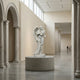 The Giant Sculptures Leaf-Winged Angel Marble Outdoor Sculpture (170cm) stands on a round pedestal in a spacious, modern museum hall with arched doorways. Two people admire the elegant statue from a distance.