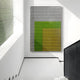 A minimalist, sunlit hallway with white walls, a staircase on the right, and Giant Sculptures’ Pixelate Lime Gold Acrylic 3D Wall Art—featuring a green-yellow gradient with a black grid—hanging on the wall.