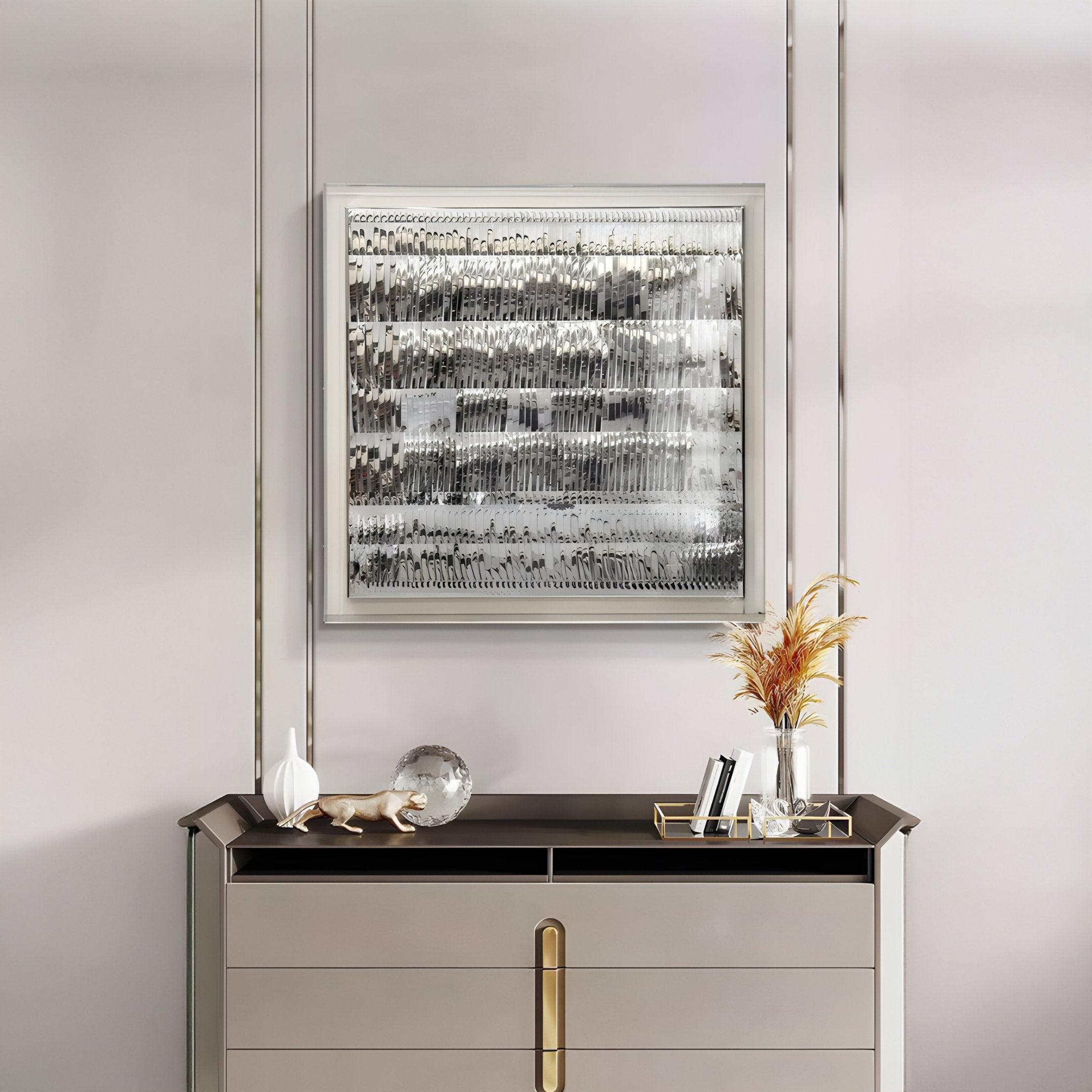 A modern sideboard with gold accents holds a glass orb, decorative animal figure, vase with dried grasses, and books. Above it hangs the Rhythm II Silver Layered Stripe Leather 3D Wall Art by Giant Sculptures on a light-toned wall.