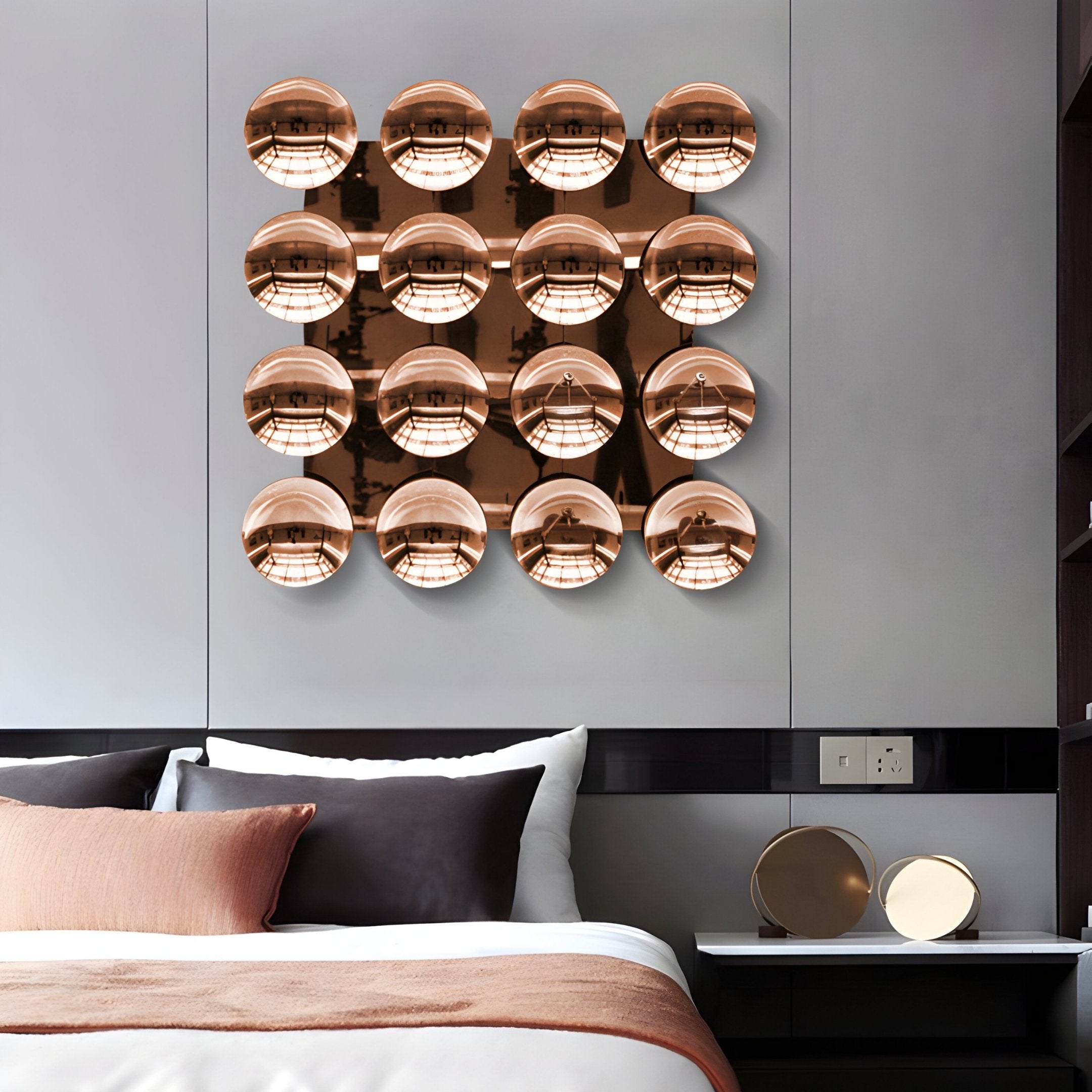 A modern bedroom features a white bed with brown and white bedding and a sculptural installation of 12 Dome Burnished Copper Acrylic 3D Wall Art pieces by Giant Sculptures arranged in a grid above the headboard. A small table holds two round mirrors.