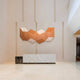 A minimalist lobby features a white marble reception desk and Giant Sculptures Vertex Orange & Cream Ridge Acrylic & Wood Carving 3D Wall Art, adding warm tones and depth against a light wall with vertical accents.