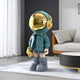 The Galactic Blue Astronaut Beats Sculpture by Giant Sculptures, standing at 100cm with a golden helmet and bold teal jacket, adorns a modern living room. Its exaggerated boots and small bag add character. A white sofa, orange chair, and draped white curtains complete the setting.