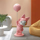 The Giant Sculptures Candy Pink Astronaut Bunny Moon Balloon Sculpture (100cm) stands on a round base in a modern room. Nearby, a curvy white table with plants and an orange chair rest on wooden floors, creating space-themed décor with whimsical charm.