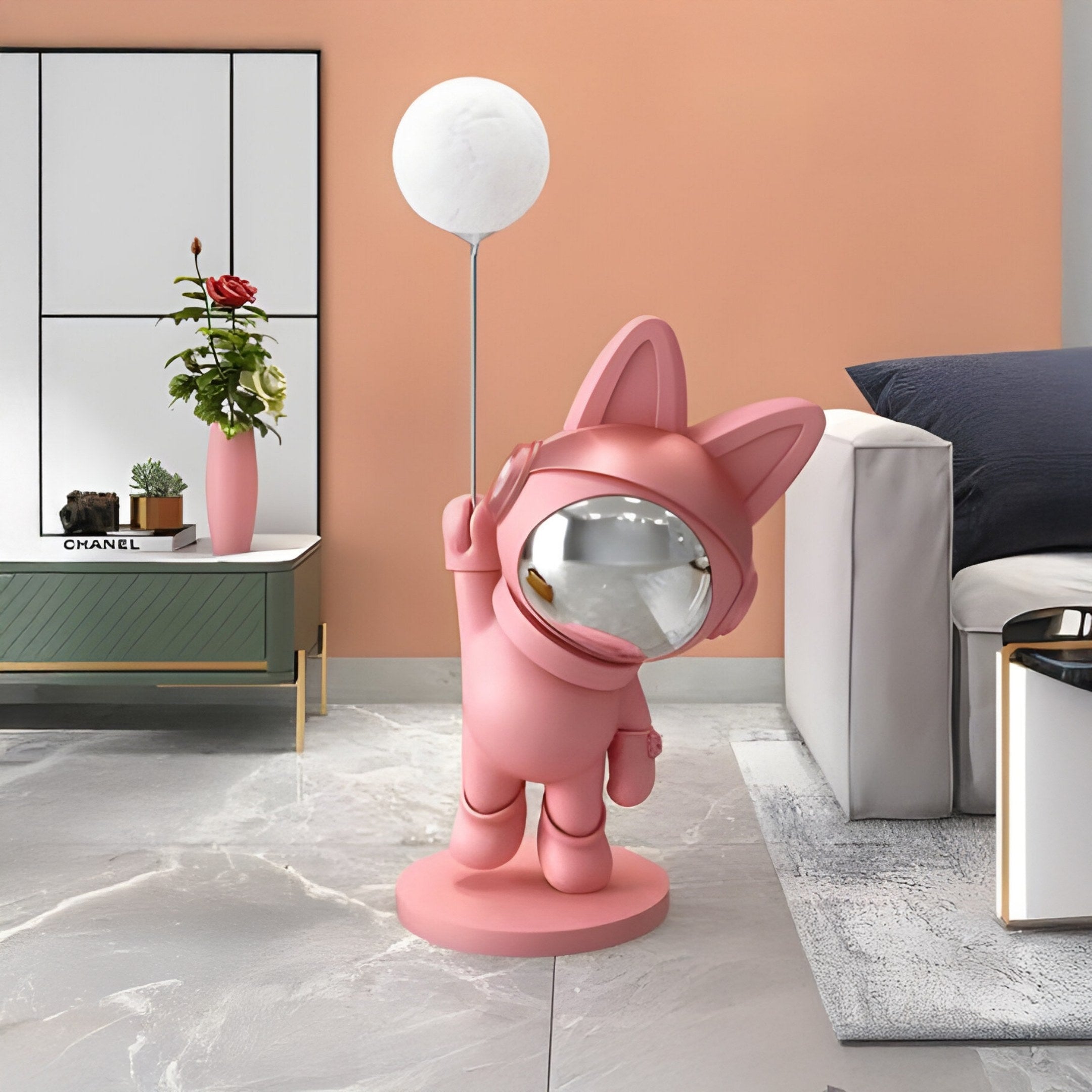 The 100cm Candy Pink Astronaut Bunny Glow Balloon Sculpture by Giant Sculptures stands on a gray marble floor near a white and gray sofa. In the backdrop, cabinets feature a vase with red flowers, while the peach-colored wall adds warmth to this whimsical scene.