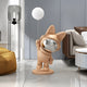 A modern living room features a sleek marble floor and a cozy brown bean bag. A stylish grey couch sits beneath the warm glow of a decorative lamp, while the Rose Gold Astronaut Bunny Glow Balloon Sculpture by Giant Sculptures adds a touch of elegance and abstraction.