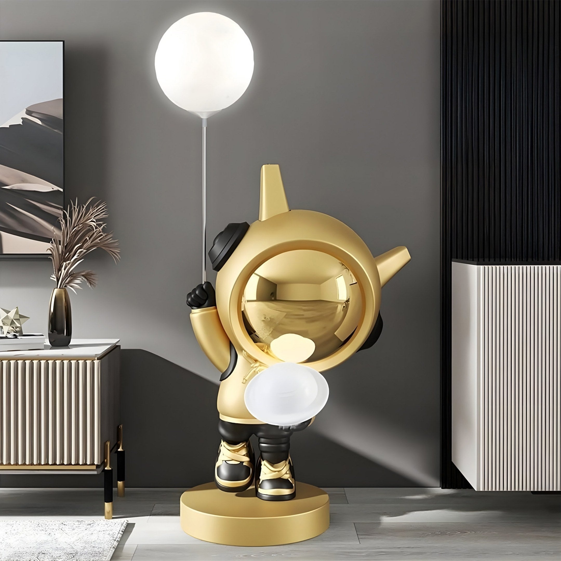 A stylish room highlights a Celestial Gold Astronaut DJ Glow Balloon Sculpture - 100cm by Giant Sculptures, with a glowing white balloon on a round base against a dark wall. Modern cabinets and vases with dried plants add to its chic, futuristic ambiance, enhanced by four-color lighting.