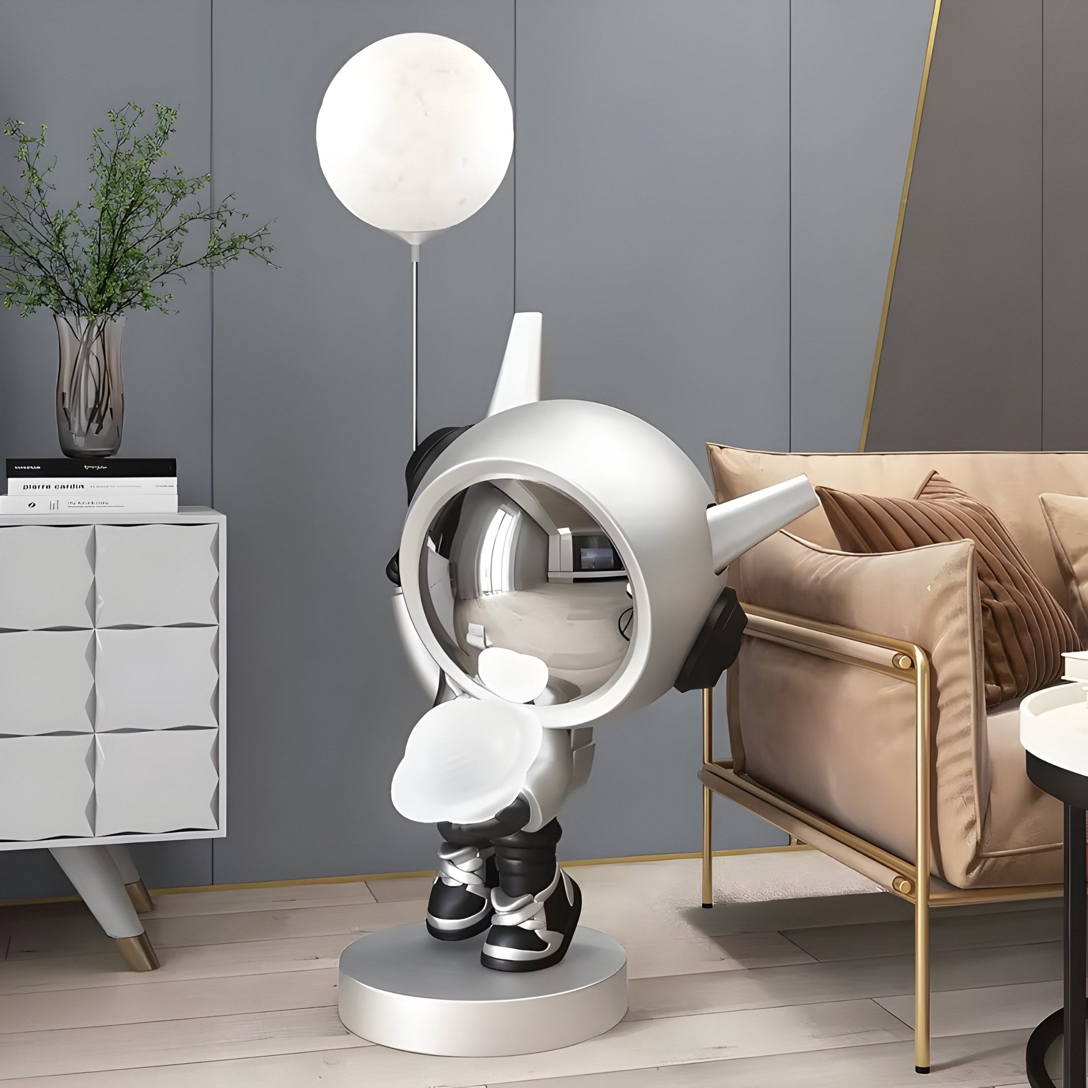 The Cosmic Chrome Astronaut DJ Glow Balloon Sculpture - 100cm by Giant Sculptures resembles a metallic astronaut holding a luminous white balloon. It complements a beige sofa, next to a small white sculpture and plant on a cabinet, set in futuristic decor.