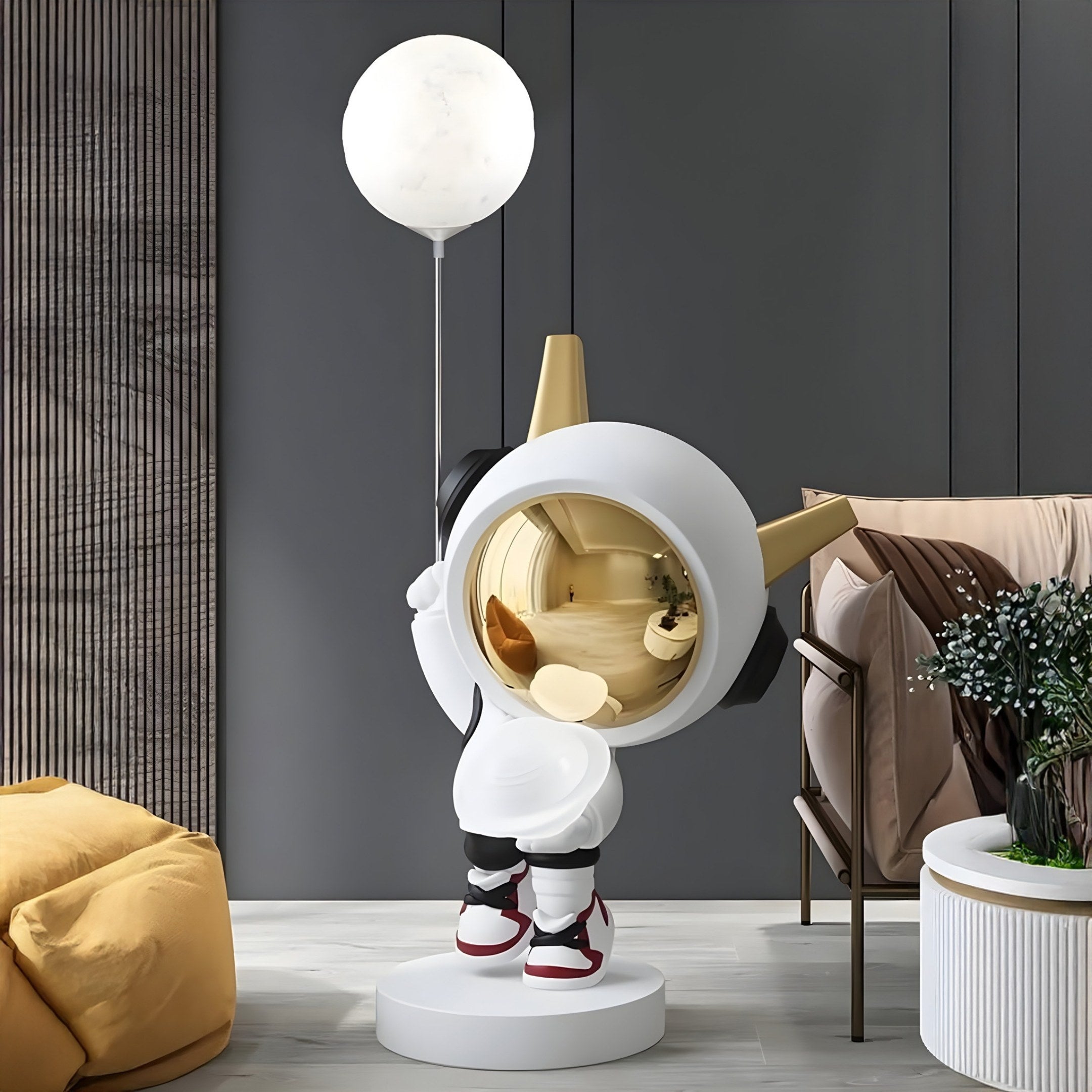 A contemporary living room features the Nova White Astronaut DJ Glow Balloon Sculpture by Giant Sculptures. The 100cm figure, resembling an astronaut with a gold helmet and glowing orb, stands on a round base near a brown chair, white table, and mustard-yellow cushion.