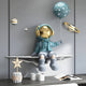 The Blue 3D Dreamer Balloon Astronaut Wall Art by Giant Sculptures showcases a resin astronaut with a gold visor holding a polka-dotted blue balloon, set against a cosmic backdrop with planet models. Nearby sit two cups, a star-shaped board, and a container of balls.