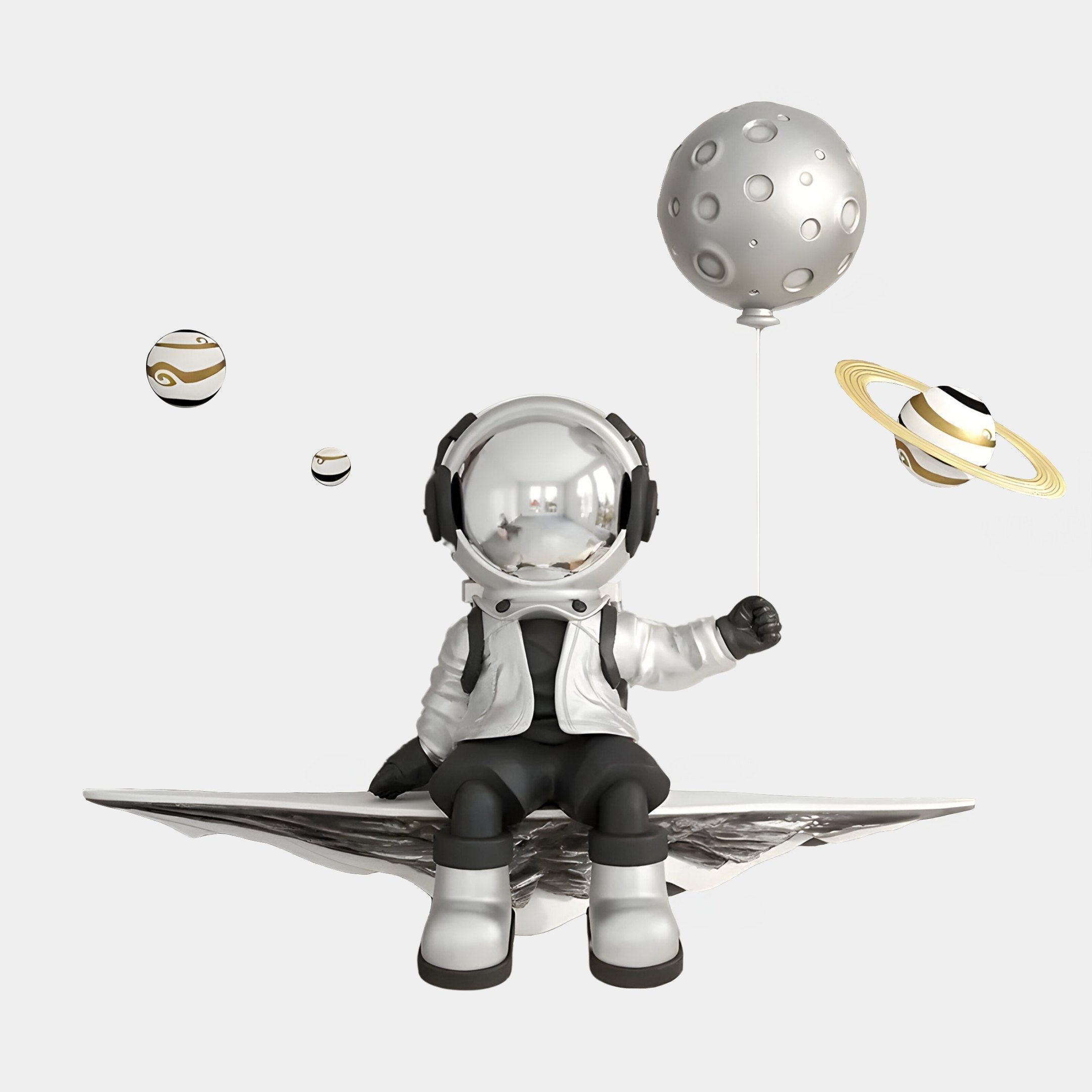 A small astronaut in a spacesuit sits on a floating platform, holding a gray planet-shaped balloon. Giant Sculptures Silver 3D Dreamer Balloon Astronaut Wall Art includes space-themed accents with three small nearby planets, one with rings, set against a white background.