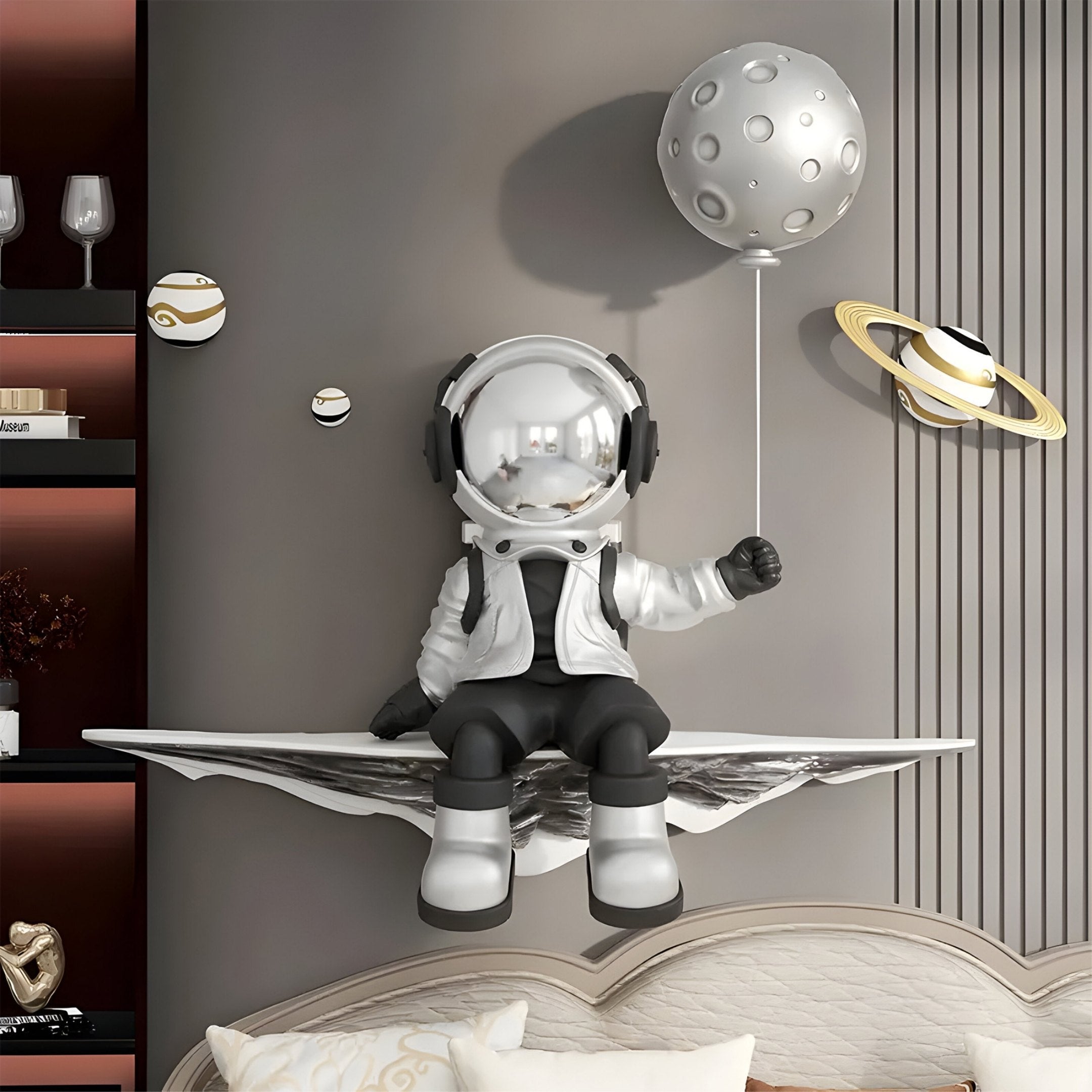 A decorative figure from the Giant Sculptures Silver 3D Dreamer Balloon Astronaut Wall Art sits on a living room shelf, holding a moon-shaped balloon with small planets. The setting includes space-themed accents and shelves of glasses and decorations.