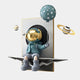 The Blue Balloon Astronaut 3D Portal Wall Art by Giant Sculptures features an adorable astronaut with a gold helmet sitting on a book edge, holding a blue balloon with white dots. Surrounding miniature ringed planets enhance the whimsical cosmic décor and space theme.