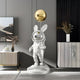 The Chrome Astronaut Floppy Bunny Balloon Sculpture by Giant Sculptures, a 106cm piece with bunny ears and a staff with a golden orb, is on display on its white platform, nestled between a modern sideboard and an abstract art piece in this stylishly modern room.