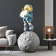 A stylish living room showcases the Galactic Blue Astronaut Glow Moonwalk Sculpture - 110cm by Giant Sculptures, depicting a cartoon astronaut with a gold helmet holding an orb on a moon replica. It includes a beige couch, modern lamp with round bulb, and marble columns.