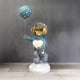 The Galactic Blue Astronaut Glow Heart Balloon Sculpture - 110cm by Giant Sculptures catches the eye with its shiny gold helmet on a blue-suited child cradling a glowing heart-shaped object resembling a USB charging lamp, accompanied by a matching blue balloon against a textured gray wall.