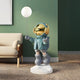 The Galactic Blue Astronaut Glow Balloon Sculpture by Giant Sculptures features a whimsical teal jacket, large boots, and a shiny gold-toned helmet that reflects light. It stands on a white base against a green wall next to a beige couch, creating an almost glow-like effect in the room.