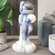 Cosmic Sky Astronaut Glow Lift-Off Sculpture by Giant Sculptures, 110cm resin figure in soft blue rising from a white cloud base while holding a glowing orb, shown in a living room beside a sofa and potted plant.