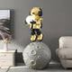 In a modern living room, the Celestial Gold Astronaut Glow Moonwalk Sculpture by Giant Sculptures stands 110cm tall on a moon-shaped pedestal, casting a soft glow like a USB charging lamp. It complements the gray wall, white cabinet with decor items, and nearby white sofa.