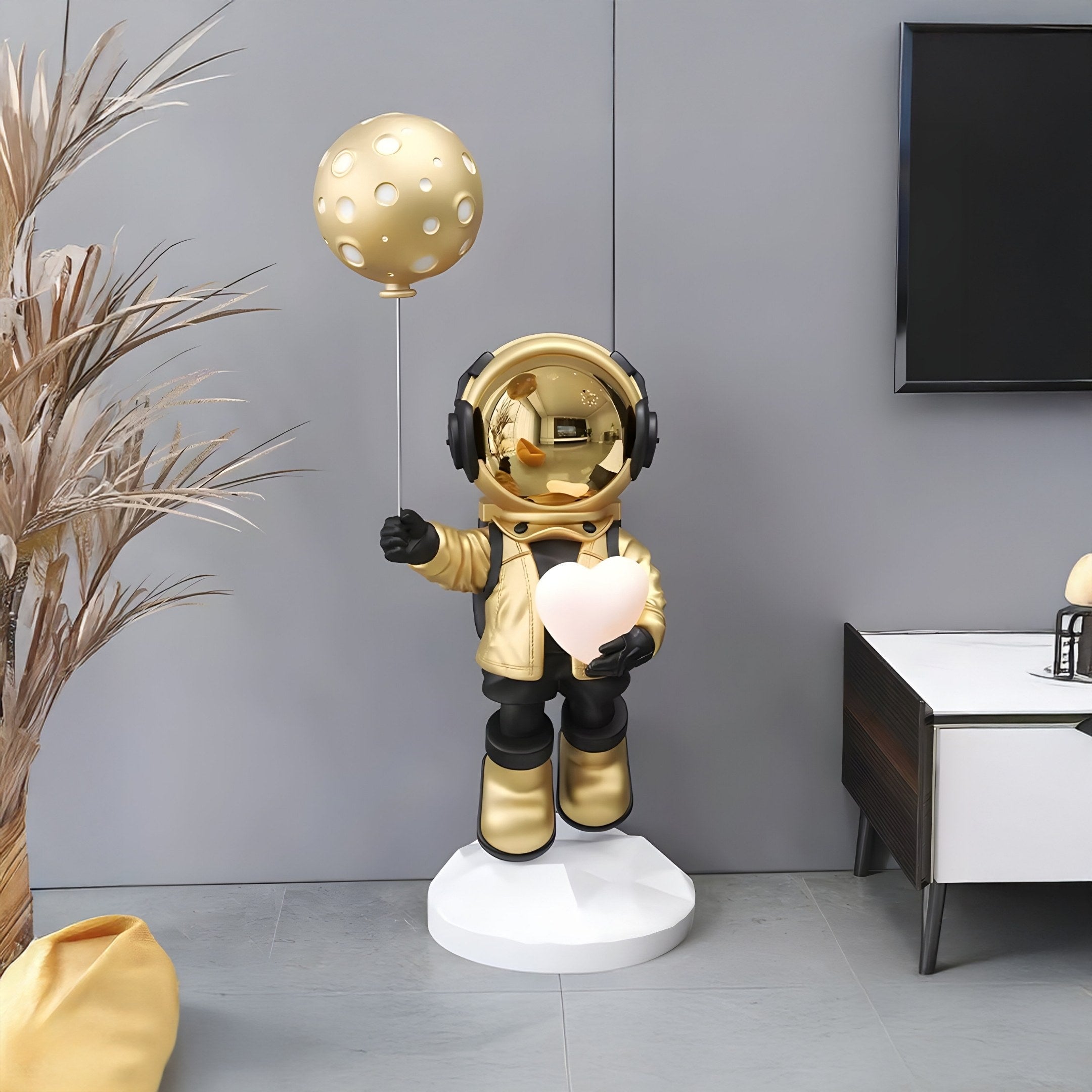 The modern room features a Celestial Gold Astronaut Glow Heart Balloon Sculpture by Giant Sculptures. The 110cm golden astronaut, wearing headphones and a jacket, stands on a white base, holding a gleaming balloon and heart. Its set amid neutral furniture with a plant beside the TV for added life.