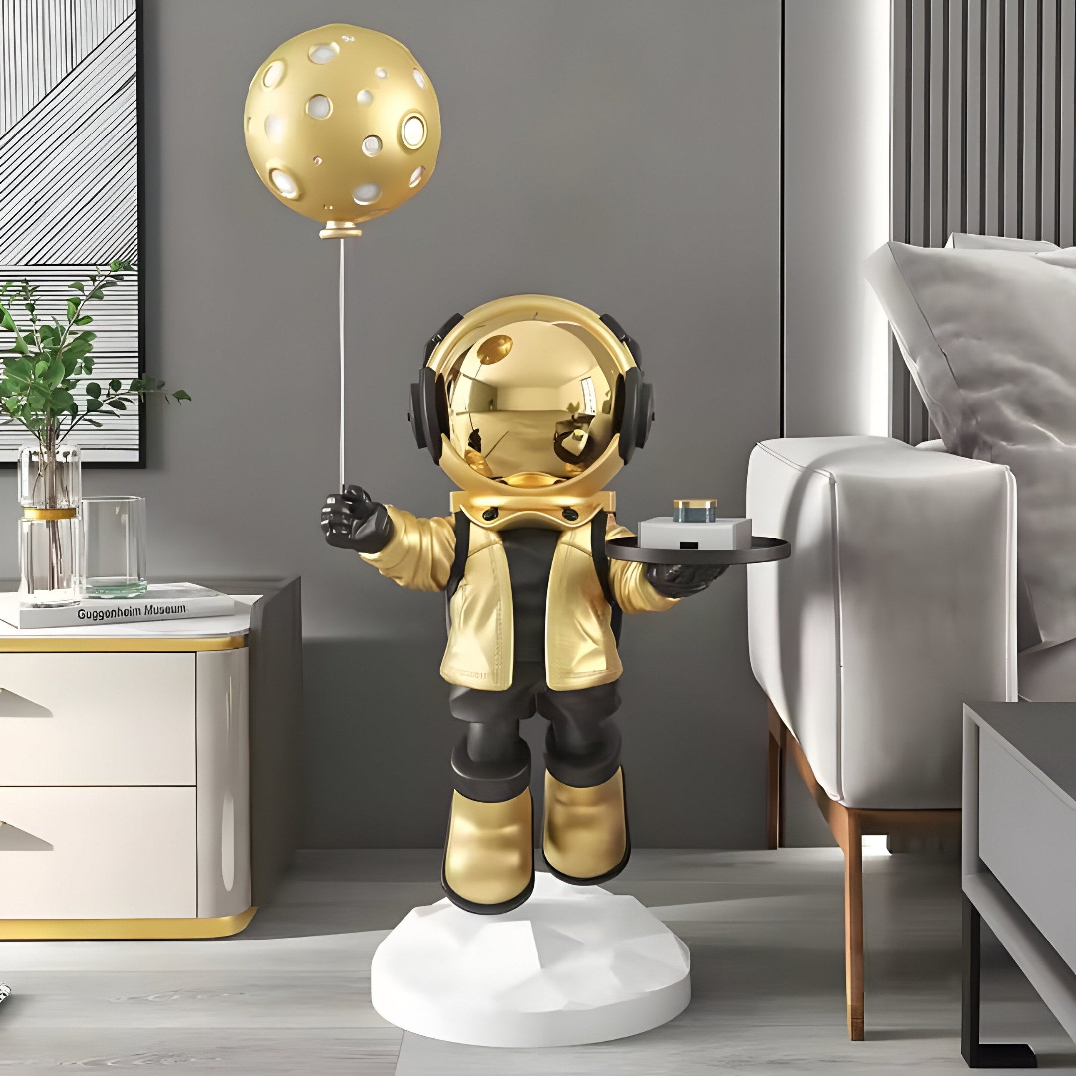 The Celestial Gold Astronaut Butler Balloon Sculpture by Giant Sculptures stands regally at 110cm on a pedestal in a modern living room, holding a tray beside the grey sofa and white cabinet adorned with books and a plant. Its reflective visor adds an elegant touch.