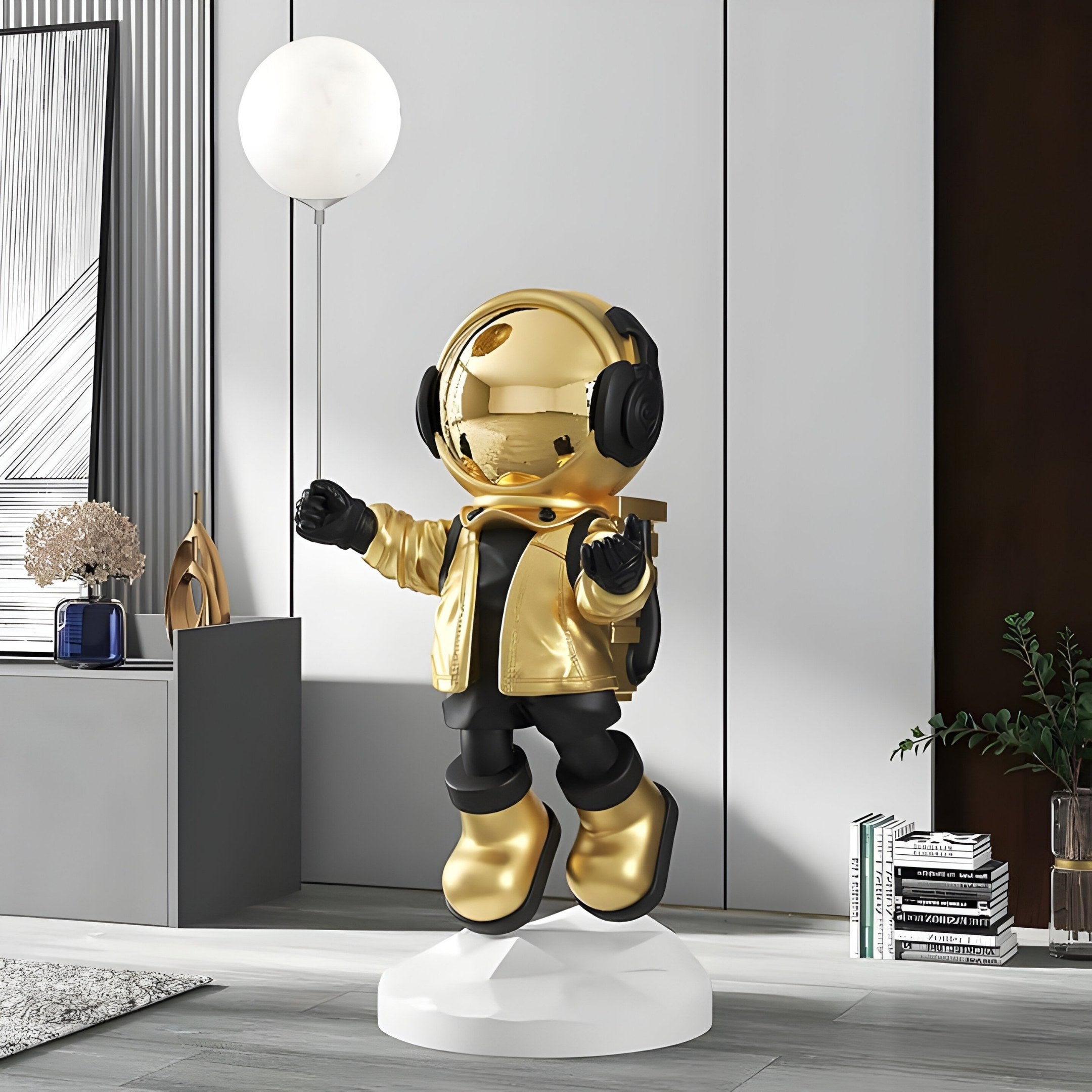 The Celestial Gold Astronaut Glow Balloon Sculpture by Giant Sculptures, standing on a white circular base with headphones and clutching a glowing balloon, beautifully complements the modern rooms grey walls and assorted decor, adding a unique space-themed charm with books and plants.